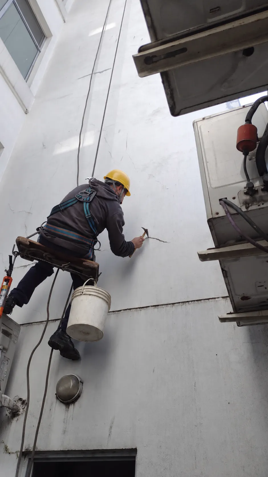 Operario sobre una silleta trabajando con herramientas en una pared de fachada.
